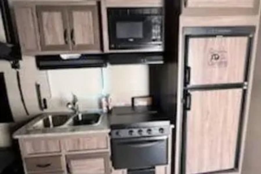 Inside an RV rental, a compact kitchen has a sink, stove, microwave, and refrigerator, next to a bed.