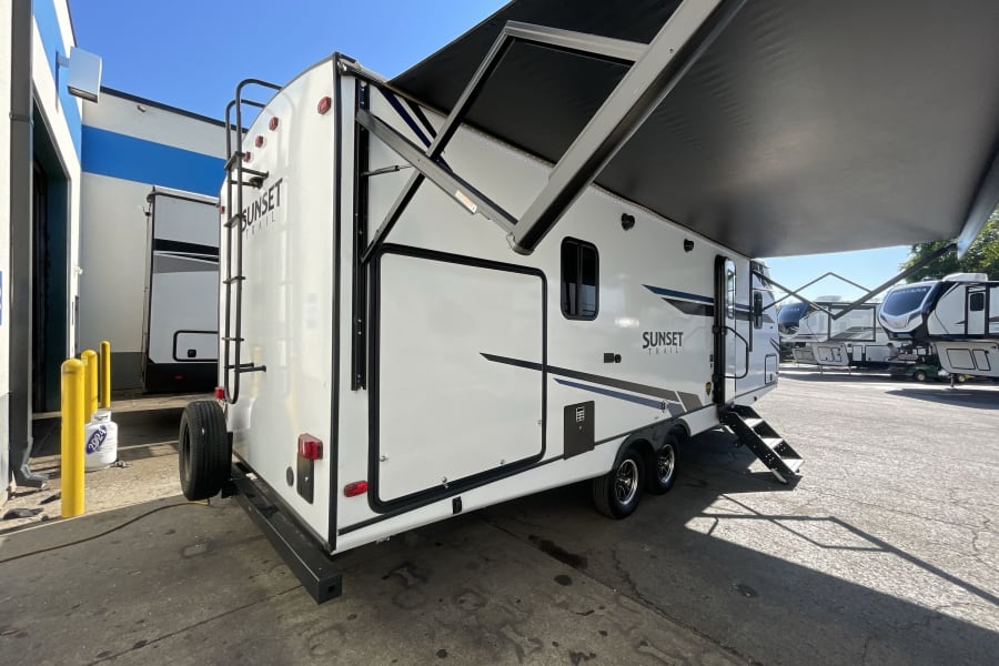A bright white Sunset Trail RV rental travel trailer with its large black awning extended, parked on a sunny asphalt lot.
