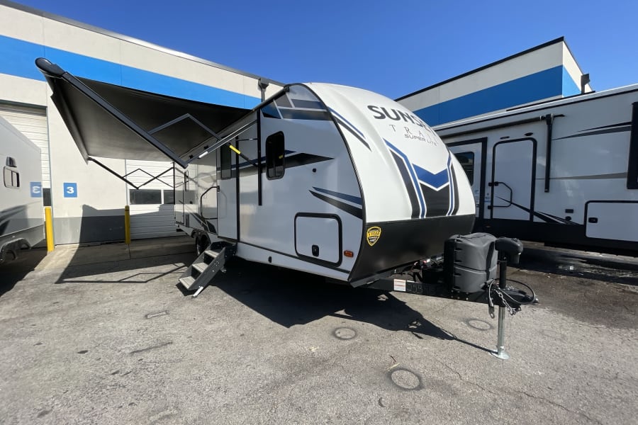 A white Sunset Trail travel trailer, an RV rental, is shown parked outdoors with its awning extended.