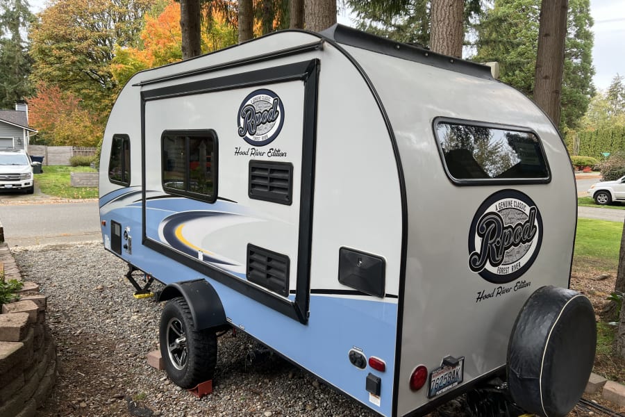 A light blue and white R-Pod camper trailer, great for an RV rental, is parked on gravel with colorful fall trees.