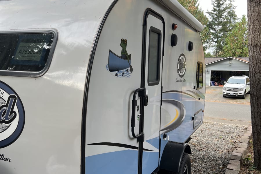 A grey and blue compact RV rental, a small travel trailer with a frog decal, is parked on gravel, ready for adventure.