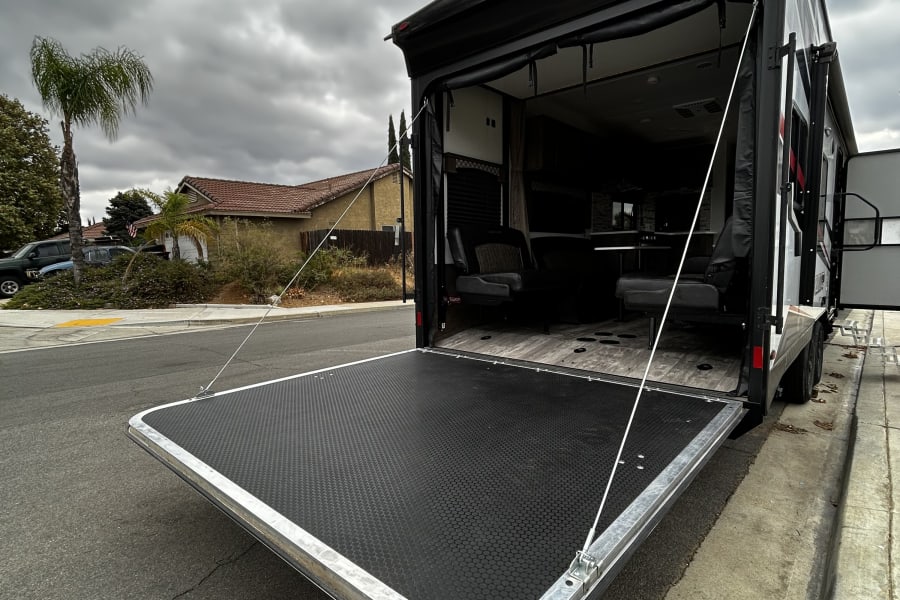 RV rental: Back of a toy hauler with its ramp lowered as a patio, parked on a residential street under an overcast sky.