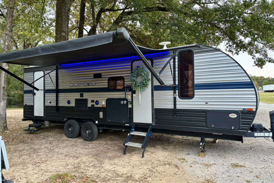 A Grey Wolf travel trailer RV rental, with a blue-lit awning and wreath, is parked at a serene campsite.