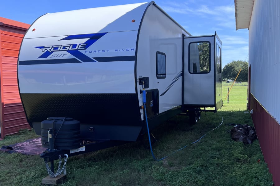 A white Forest River Rogue rv rental (travel trailer) parked on grass between red and white barns under a clear blue sky.