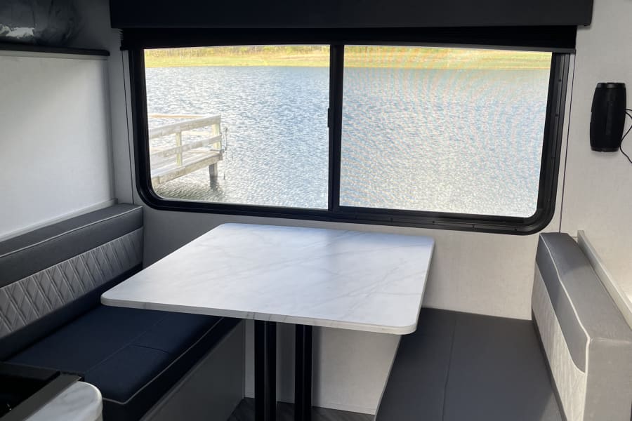 This RV rental features a modern dinette with light grey cushions, a white table, and a window offering a serene lake view.