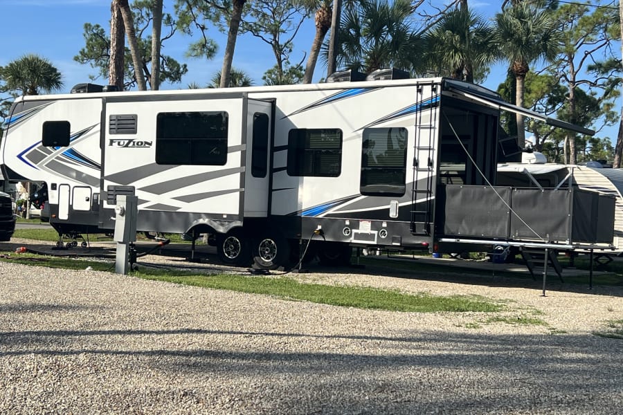 Spacious Fuzion fifth-wheel RV rental parked at a sunny campsite with palm trees & a rear patio setup.