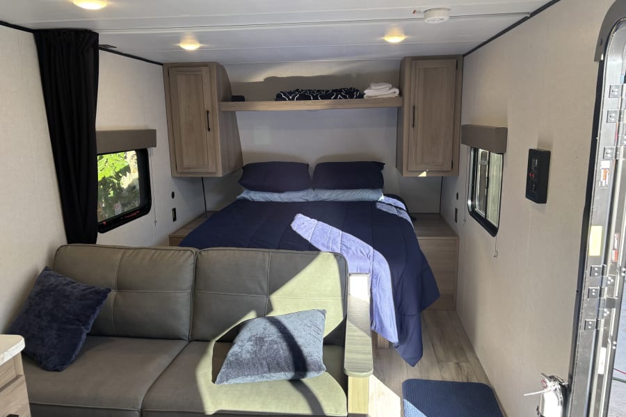 View of a modern rv rental interior, showing a cozy bed, sofa, windows, and compact living space with natural light.