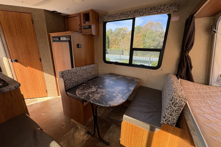 Inside an RV rental: cozy dinette area, compact kitchenette with fridge, and window overlooking trees.