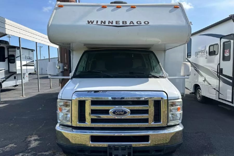 Front view of a white Winnebago Class C RV on a Ford chassis, parked at an RV rental lot with other units.