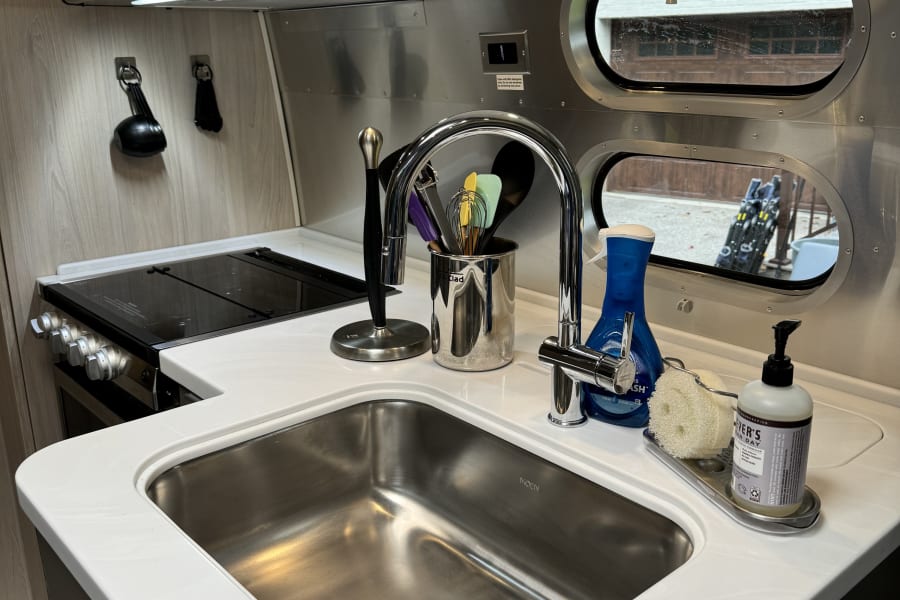 Inside a modern RV rental: a compact kitchen with a stainless steel sink, cooktop, & bright white counters.