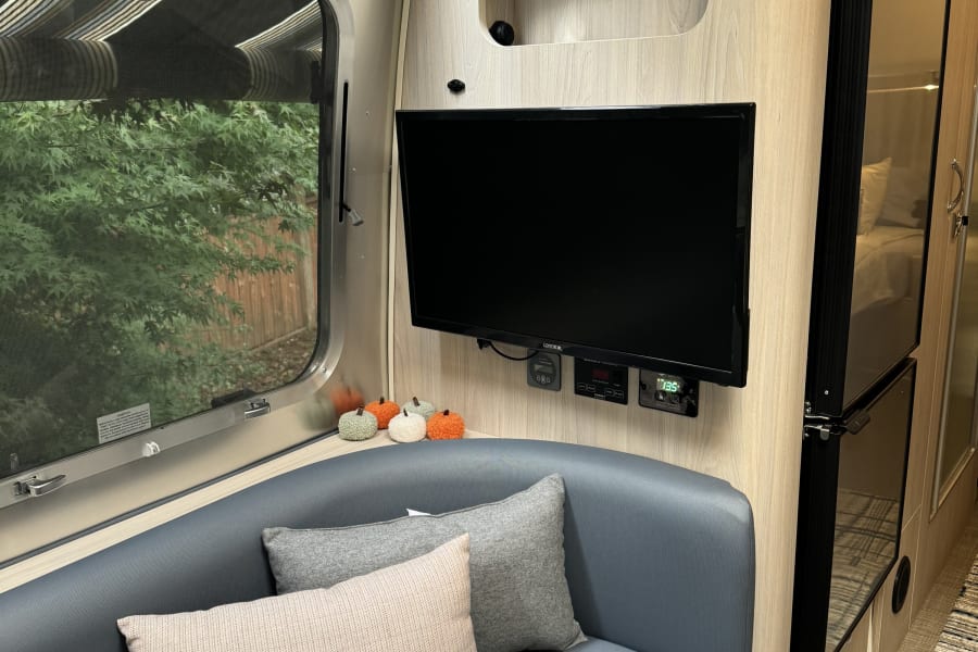 Cozy RV rental interior: A blue sofa, mounted TV, window with tree view, & fridge glimpse.