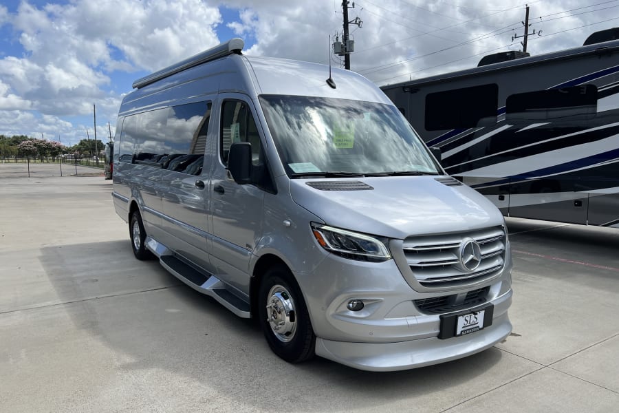 A silver Mercedes-Benz Sprinter van, suitable for RV rental, parked on a concrete lot under a blue, cloudy sky.