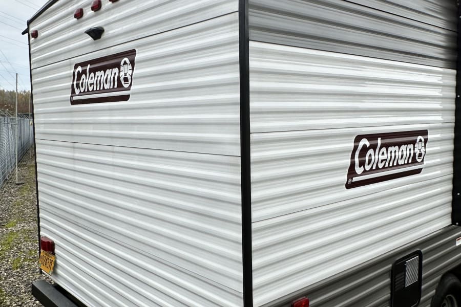 This Coleman travel trailer, ideal for your next RV rental, shows its rear and side with grey and white siding, tail lights, and an access panel.