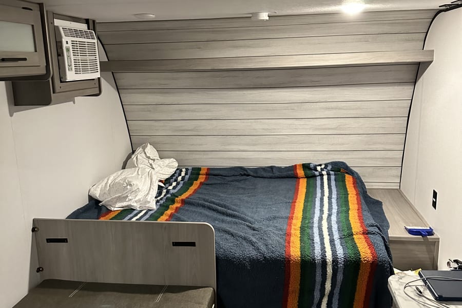 Inside a compact RV rental, featuring a comfortable bed with a vibrant blanket, an AC unit, and flexible seating.