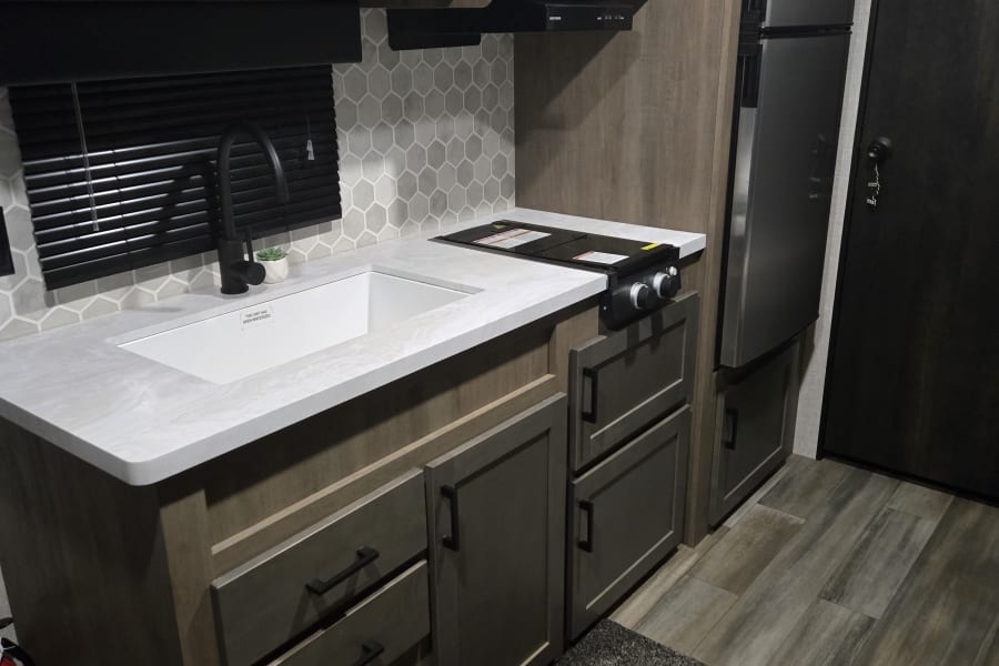 Compact rv rental kitchen: white sink, black faucet, cooktop, microwave, and stainless steel fridge with wood cabinetry.