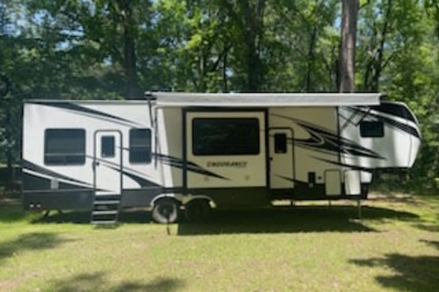 This white fifth-wheel RV rental, with awning out, is set up at a green campsite. Perfect for your next trip!