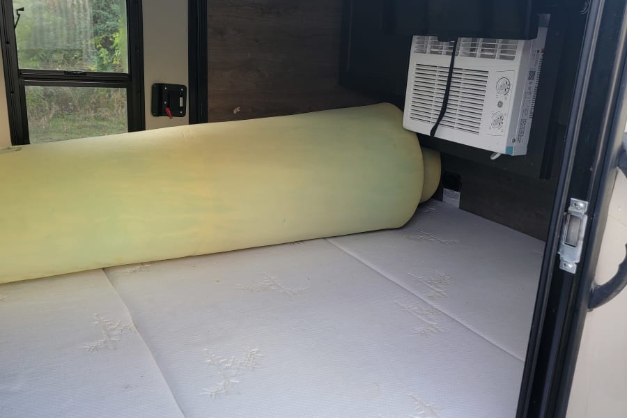 RV rental interior: A mattress with a rolled foam topper, a window AC unit, and an EXIT sign above the side window.