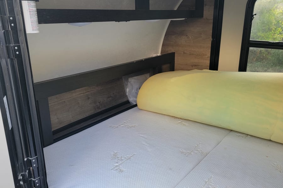 RV rental interior: Comfortable bed with yellow foam topper, overhead storage, and fire extinguisher for safety.
