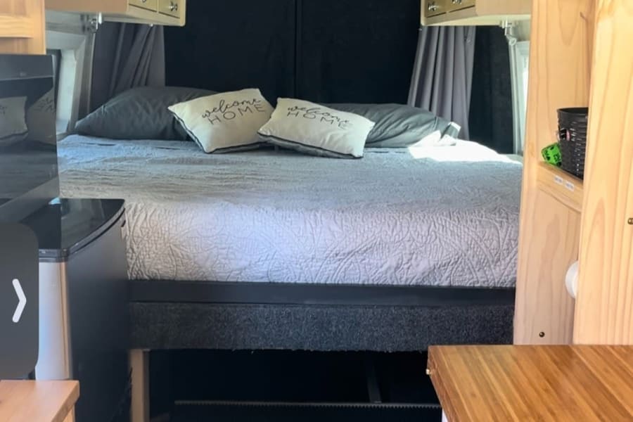 Inside this RV rental, find a cozy camper van interior with bed, overhead cabinets, and clever storage.