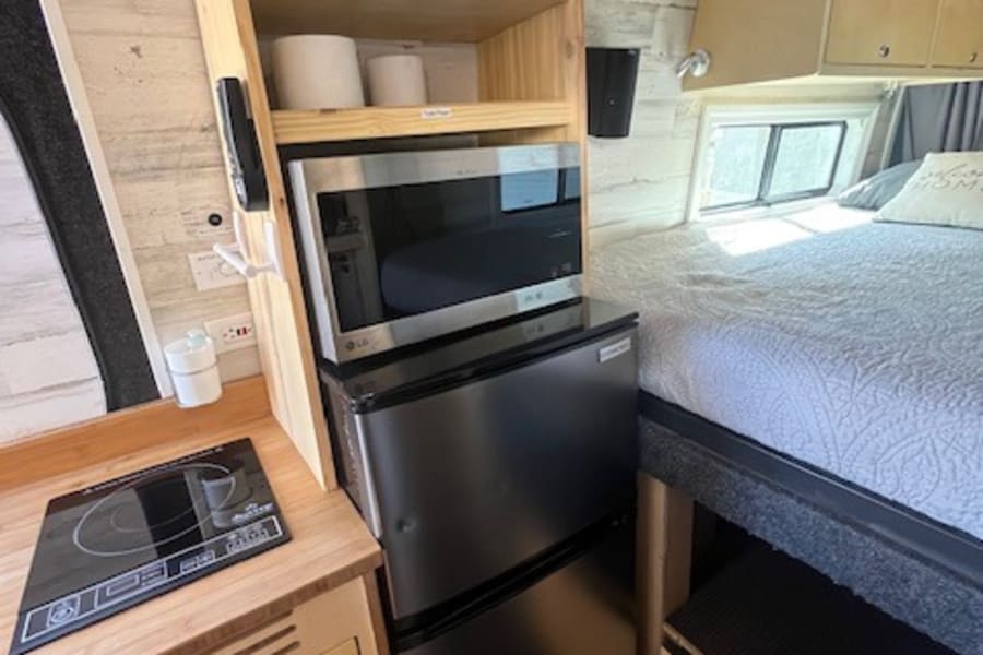 Cozy rv rental interior: compact kitchen (induction cooktop, microwave, fridge), bed, and built-in storage.