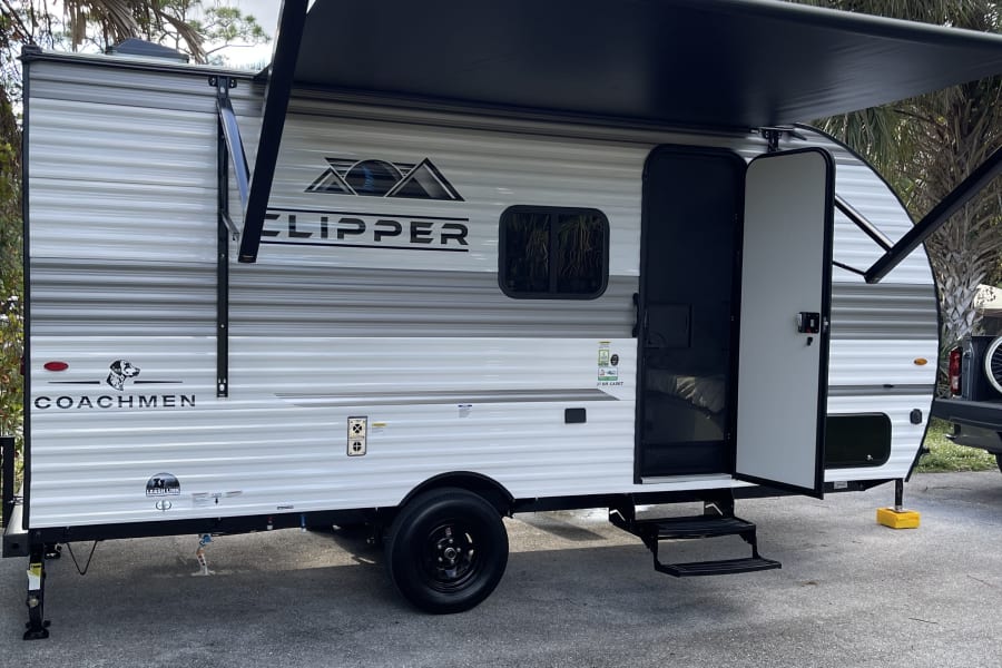 Experience nature with this white and gray Coachmen Clipper RV rental. Its black awning is extended, door open, parked on asphalt.