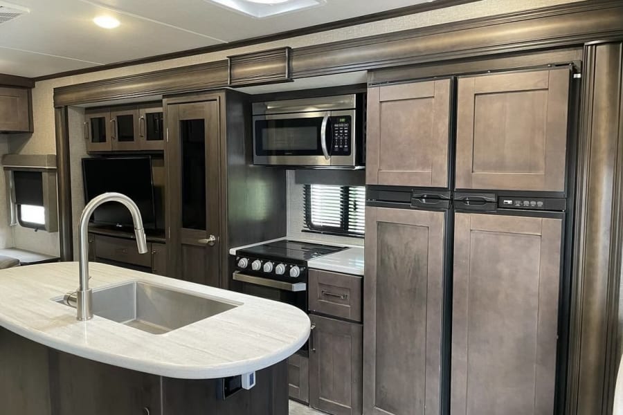 Modern RV rental interior showcasing a well-equipped kitchen island, stove, microwave, fridge, and TV living area.