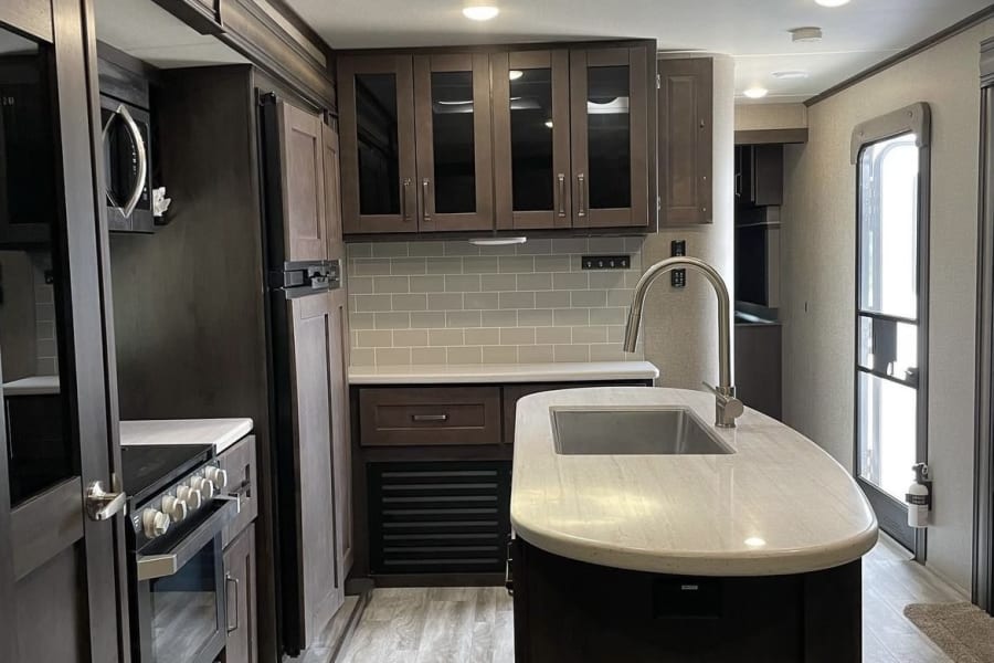 Spacious RV rental kitchen featuring dark cabinetry, light countertops, integrated appliances, and a central island with sink.