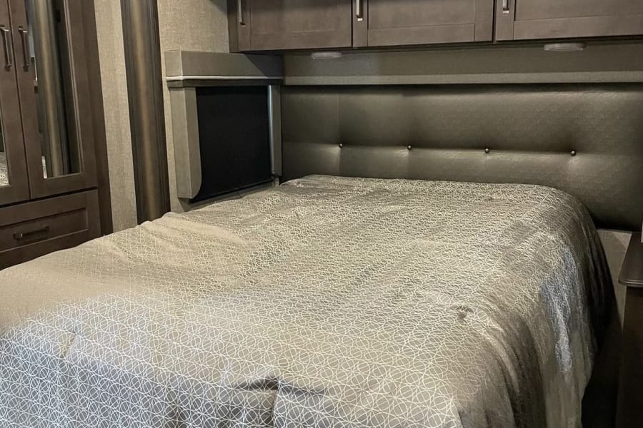 Inside an RV rental: a modern bedroom with a grey patterned bedspread, dark padded headboard, and overhead storage.