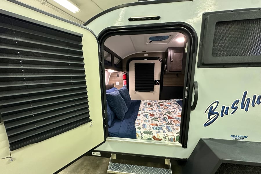 Step into this compact Bushwacker RV rental, showcasing its cozy interior bedroom with bed, storage, and open door.