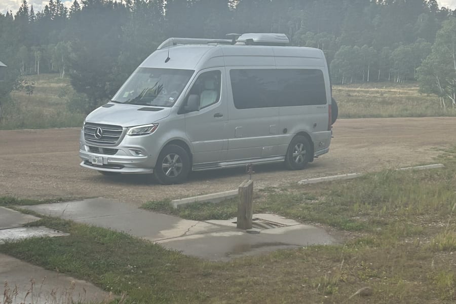 An RV rental, a silver Mercedes Sprinter camper van, parked at a rustic site surrounded by forest.