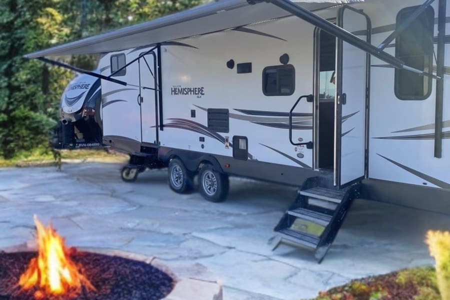 Relaxing RV rental setup with a white camper, extended awning, and a cozy fire pit on a patio amidst forest trees.