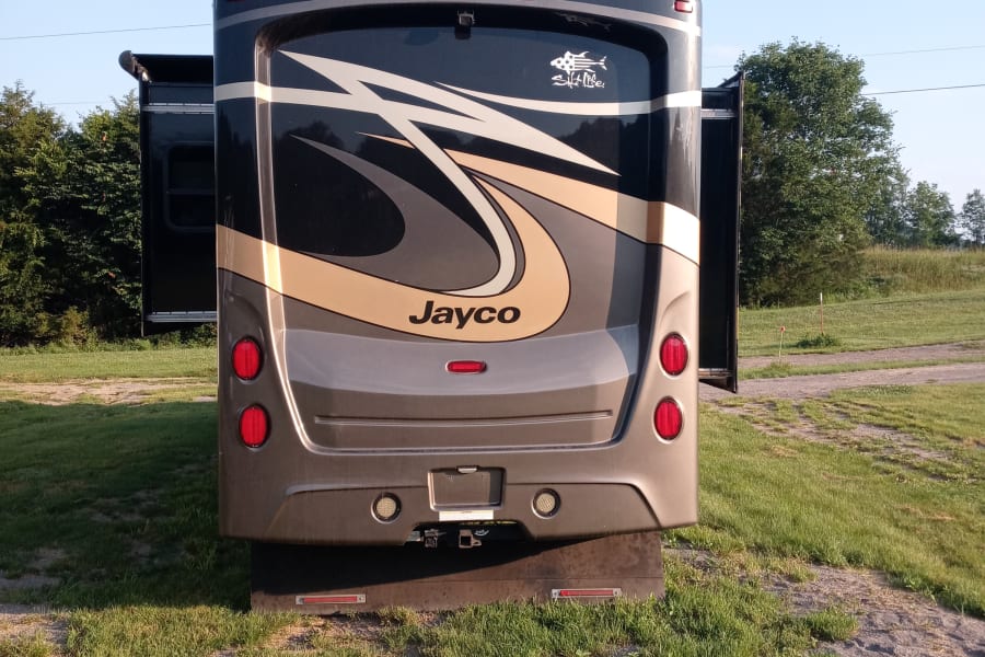 Rear of a dark Jayco RV with slide-outs extended, parked on grass/gravel. Ideal for an RV rental adventure under clear skies.