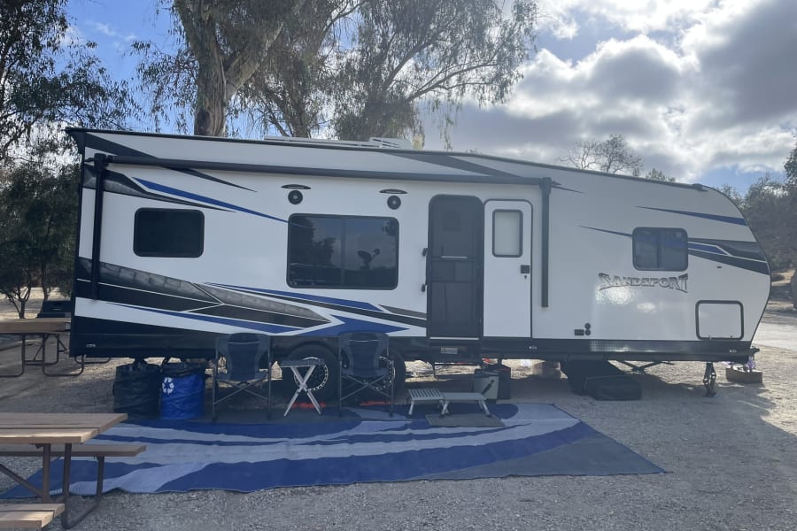 This RV rental at a sunny campsite features an outdoor mat, chairs, and picnic table, perfect for camping.