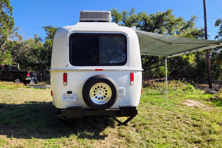 White RV rental camper with an awning extended and spare tire, parked outdoors in a grassy area under a blue sky.