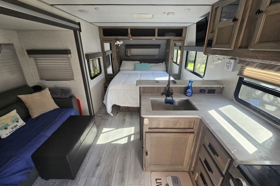 Cozy RV rental interior with bed, sofa, and kitchen sink bathed in sunlight, offering a comfortable living space.