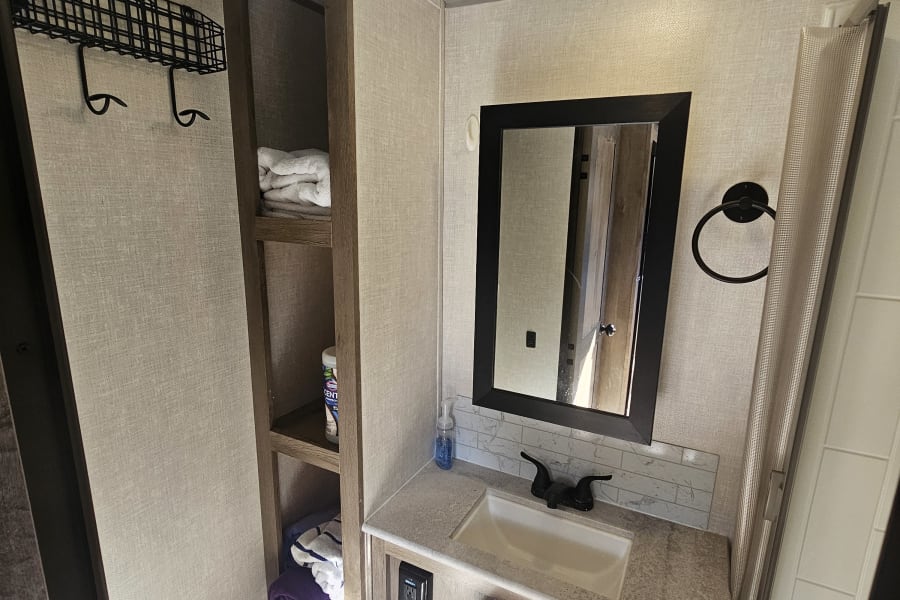 Inside an RV rental bathroom, featuring a modern sink, toilet, mirror, overhead light, and practical storage shelves.