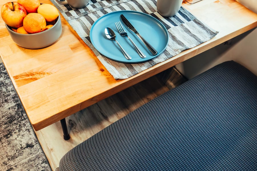 Inside an RV rental: a bright dining area featuring a wooden table with two place settings, fruit, and snacks by the window.
