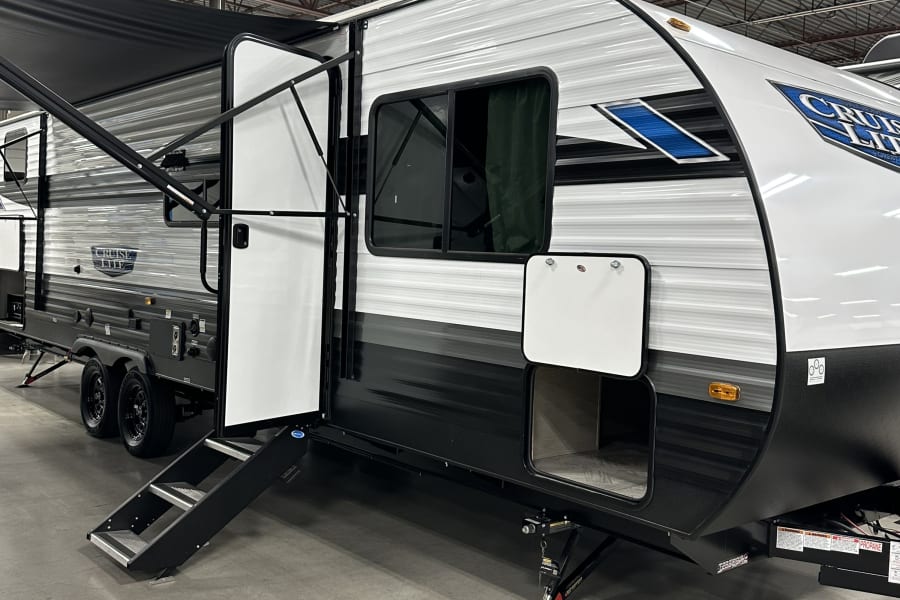 Explore travel options with this RV rental! A white & black travel trailer, featuring an open door with steps & an awning.