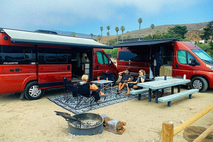 Two red RV rental vans at a sandy campsite. People relax outdoors with a picnic table, chairs, and a fire pit.