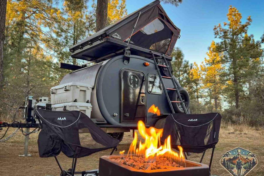 Cozy RV rental setup: a teardrop trailer with a rooftop tent, two chairs, and a warm fire pit in a wooded campsite.