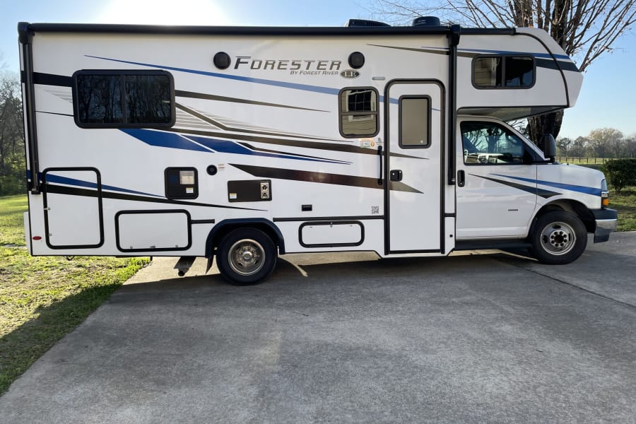 This white Forester Class C RV rental, with blue and black accents, is parked on a concrete driveway in sunlight.