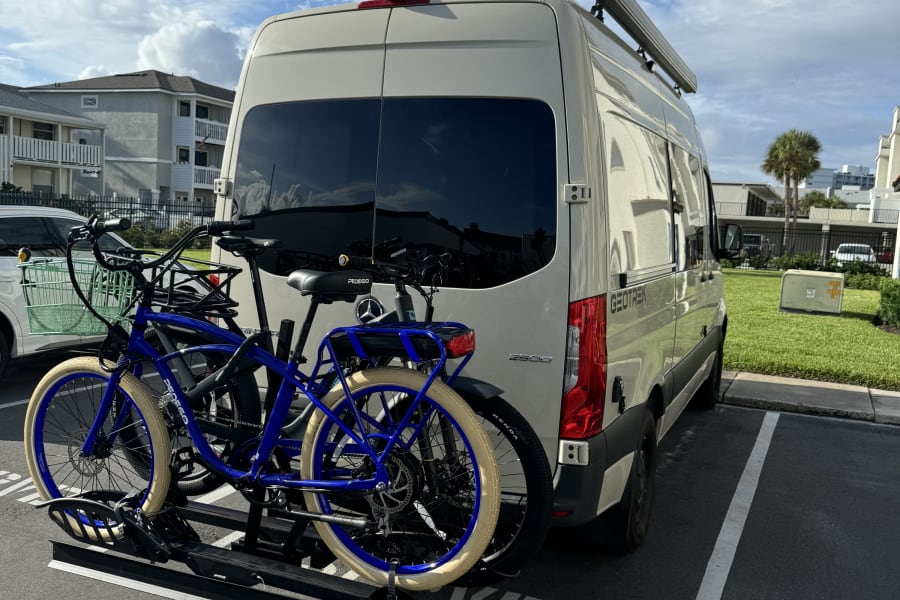 Mercedes-Benz RV rental van with two blue bikes on a rear rack, parked in a sunny lot.