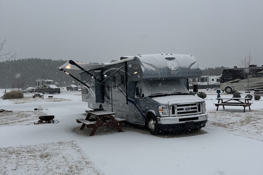 Winter wonderland at an RV park: A snow-covered gray motorhome (RV rental) with its awning extended. Picnic table and snow-dusted ground visible.