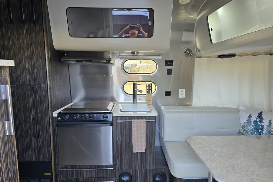 Compact RV rental kitchen & dining area. Features a stainless steel stove, sink, and light gray booth seating.