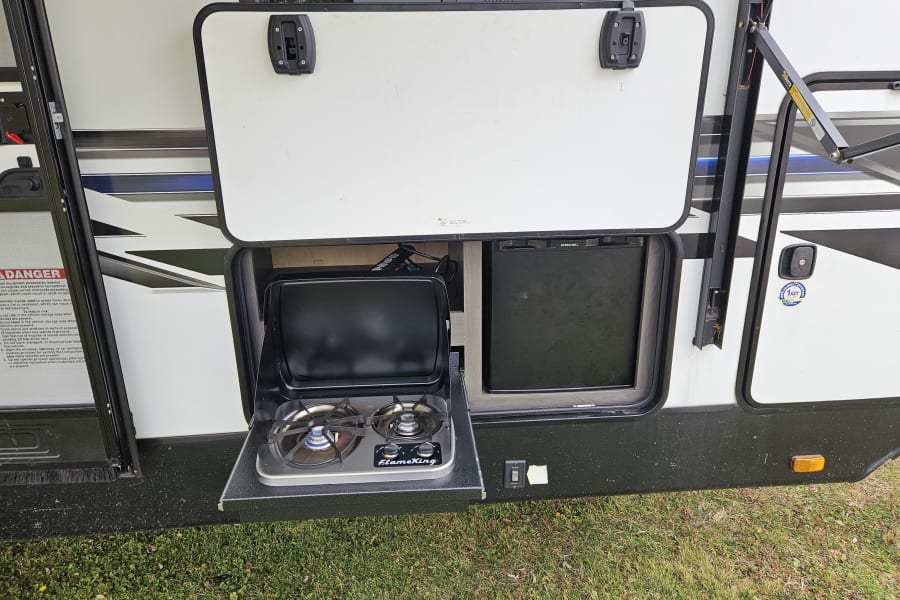 This RV rental boasts an outdoor kitchen featuring a FlameKing stove and EverChill mini-fridge, ideal for cooking outdoors.
