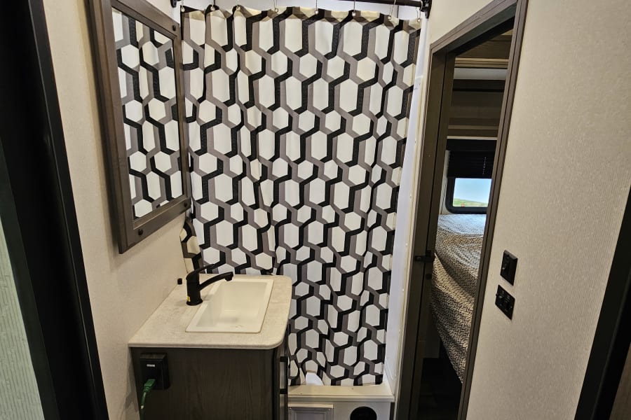 RV rental bathroom with toilet, sink, mirror, and shower. A black, white, and grey geometric curtain hangs in the shower area. Skylight visible.