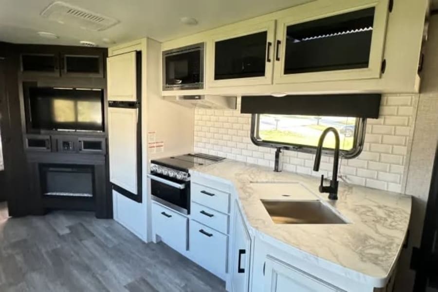 White & grey RV rental interior: modern kitchen with subway tile, sink, stove, microwave; plus TV & fireplace.