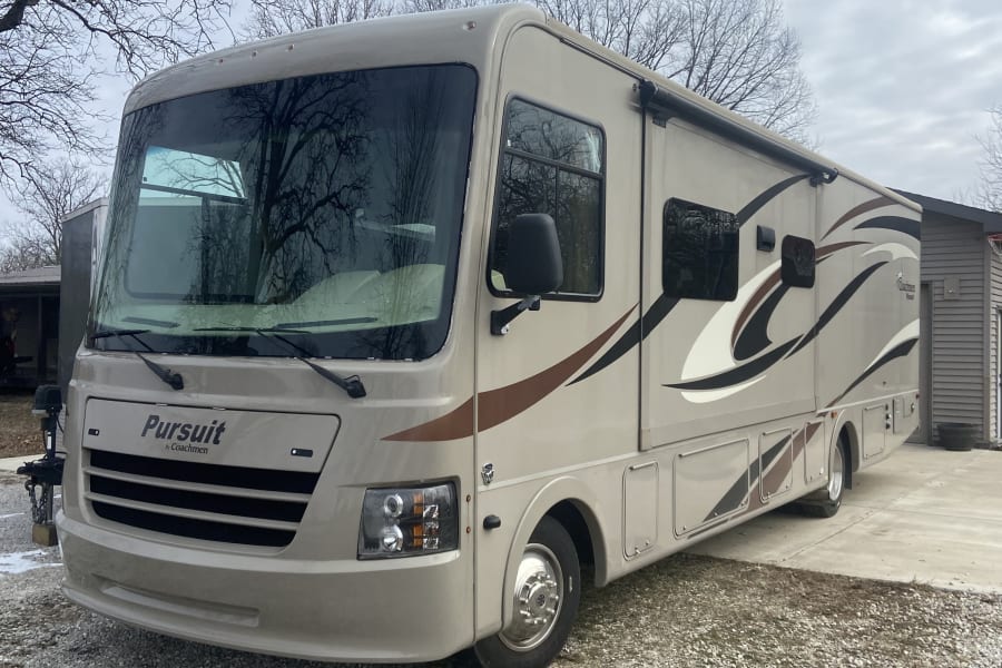A large, tan Coachmen Pursuit motorhome parked on a gravel driveway, ready for rv rental.