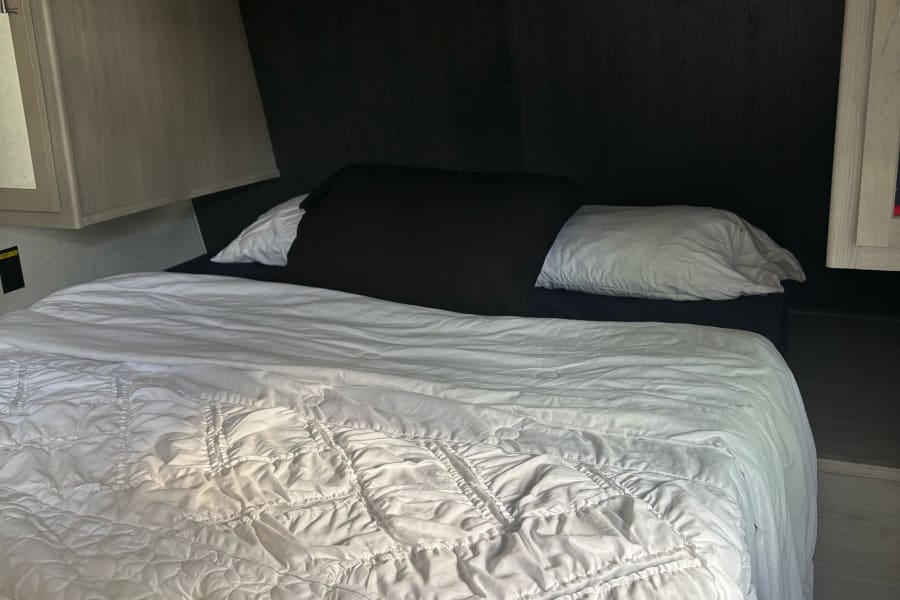 Cozy RV rental bed with white bedding, dark headboard, and wood cabinetry overhead.