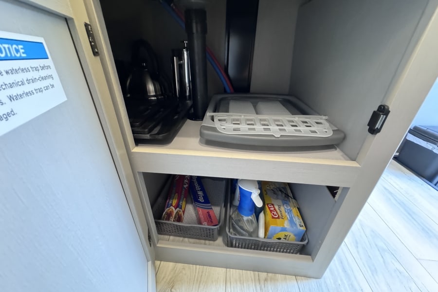 Compact under-sink storage in an rv rental, showing plumbing, a collapsible drying rack, cleaning spray, foil, and trash bags.